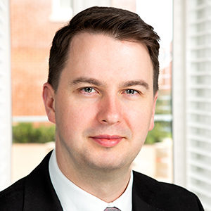 Headshot of Dominic Maher in a black suit looking into the camera